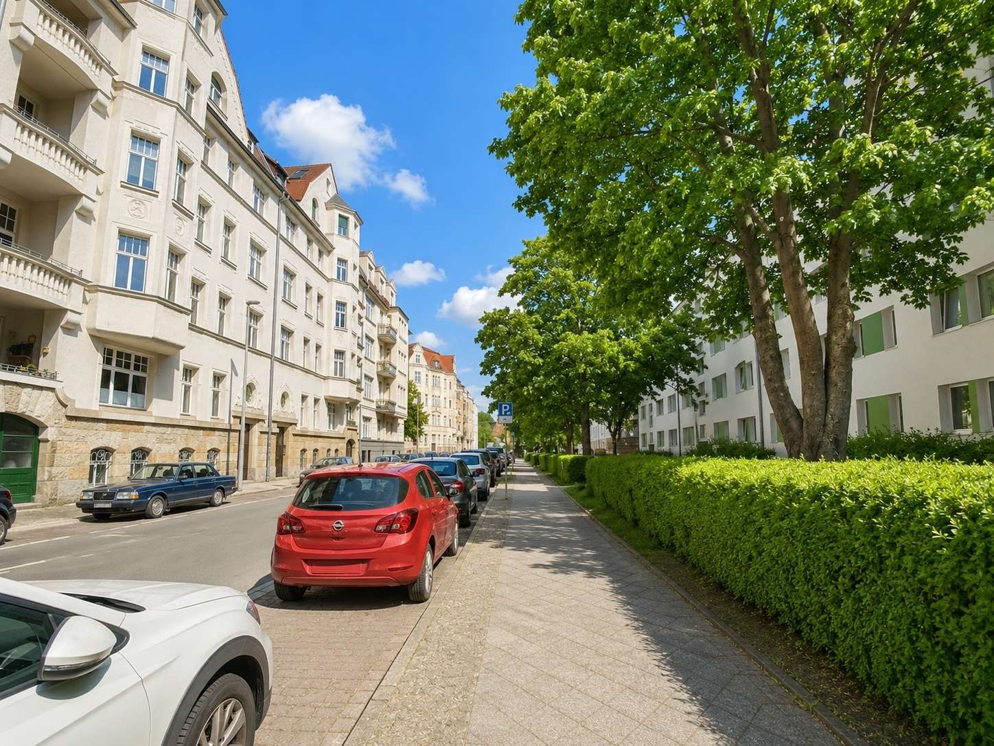 A sunny urban street with parked cars along the curb, cream apartment buildings on the left and green trees with a hedge on the right under a blue sky.