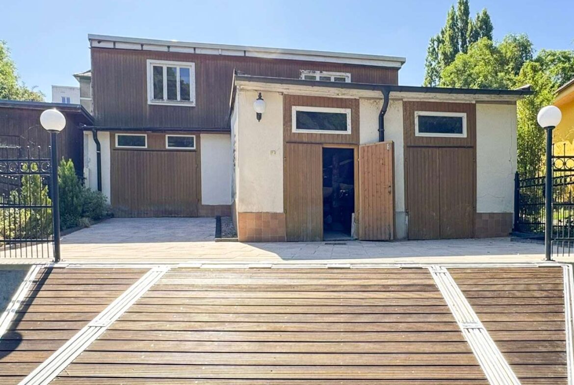 Two-story brown house with white trim and wooden doors, flanked by gates, seen from a wide wooden deck on a sunny day.