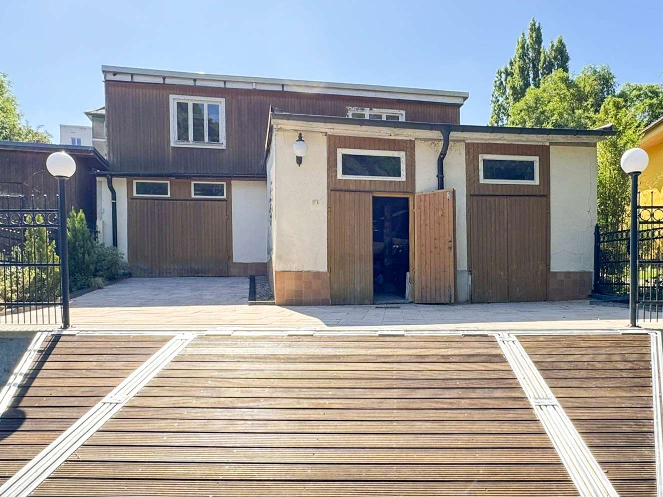 Two-story brown house with white trim and wooden doors, flanked by gates, seen from a wide wooden deck on a sunny day.