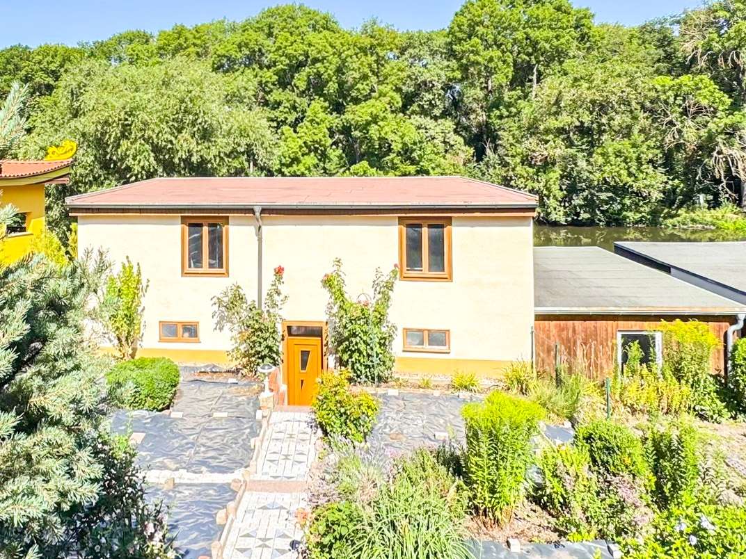 Two-story beige house with orange front door and brown-framed windows, set in a lush, landscaped garden with a stone walkway.