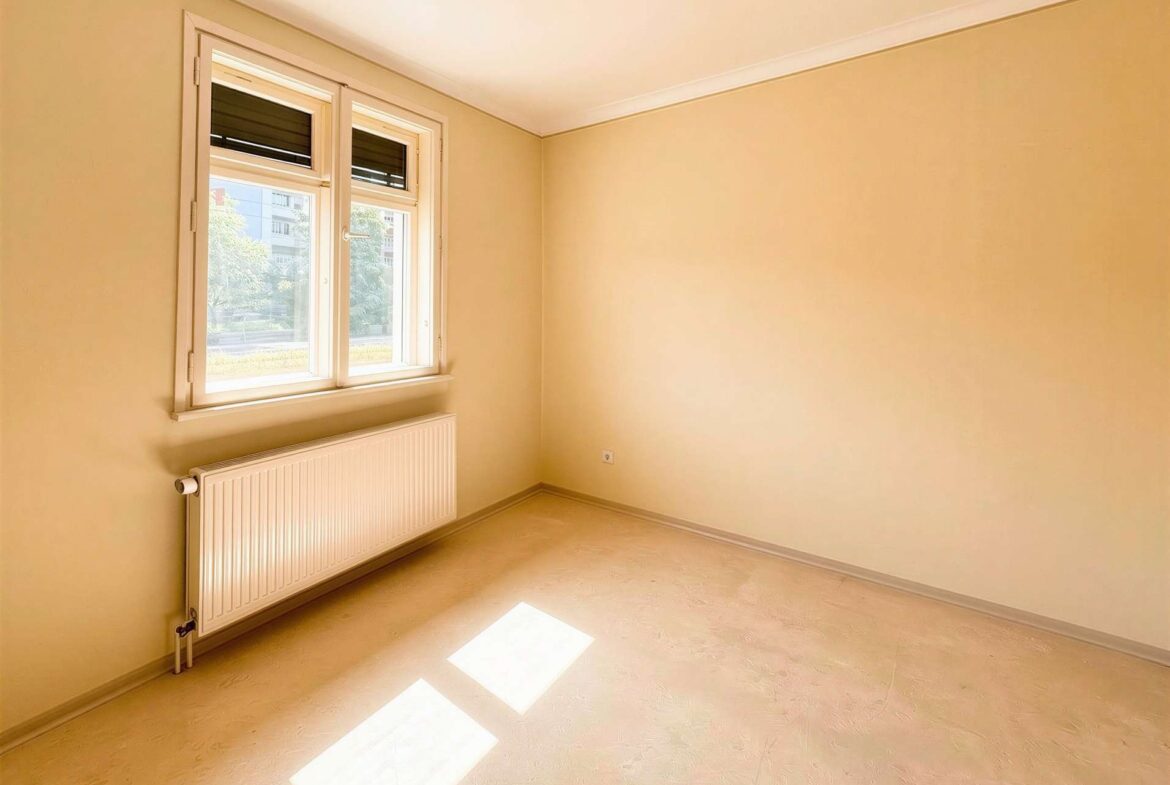 Bright empty room with cream walls, a double window and radiator beneath, sunlight casting rectangular patches on the floor