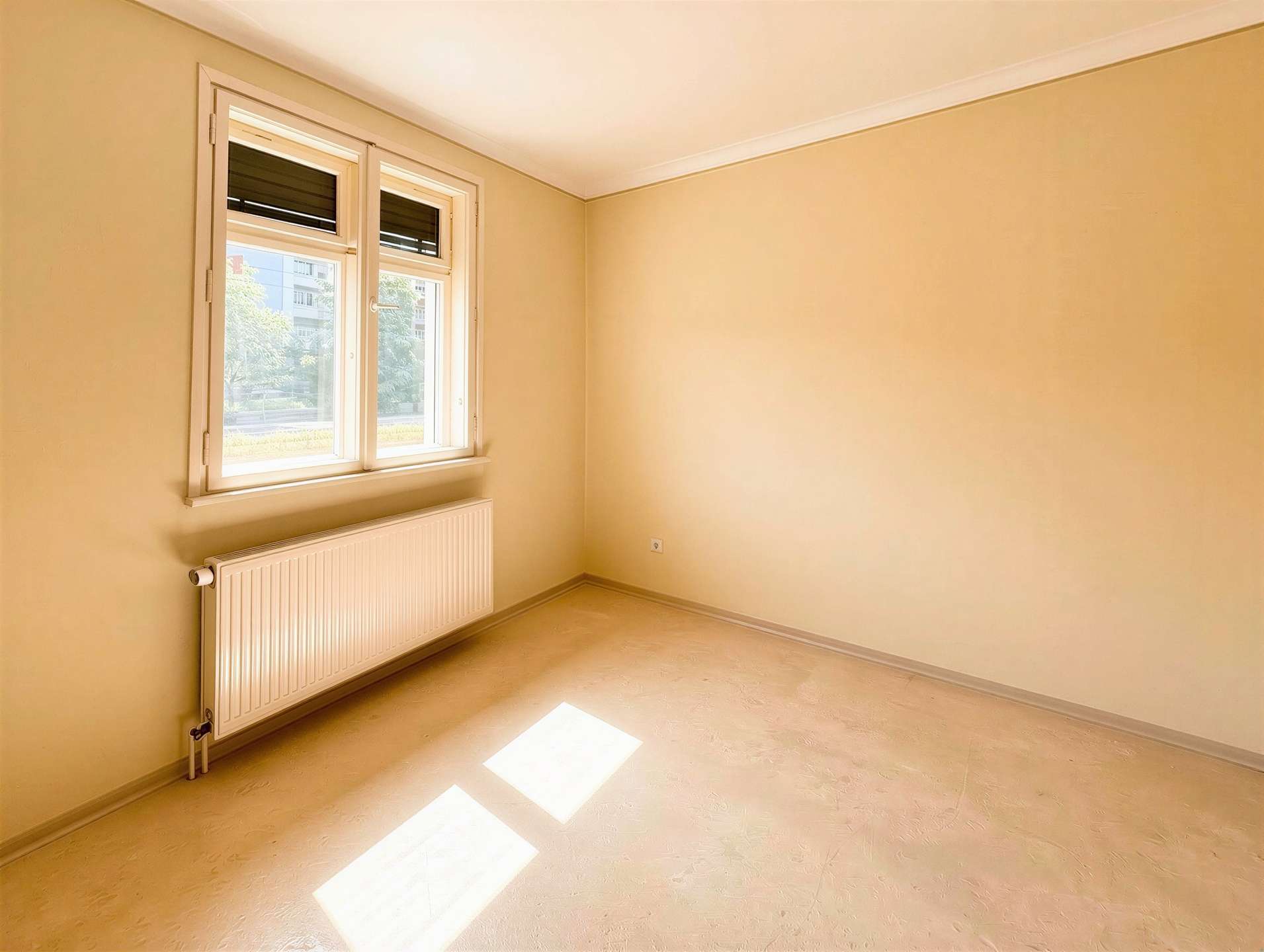 Bright empty room with cream walls, a double window and radiator beneath, sunlight casting rectangular patches on the floor