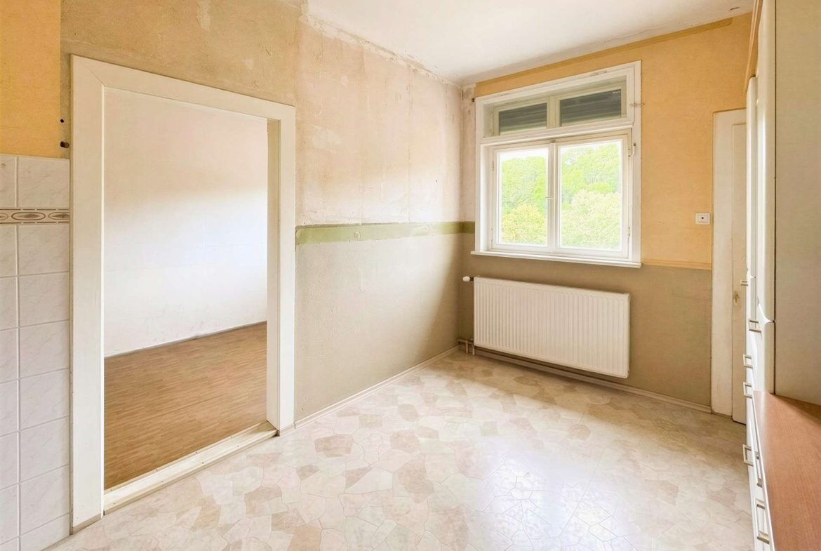 A bright, unfinished kitchen with bare plaster walls, a large window, radiator, and tiled floor; doorway to another room visible on the left.