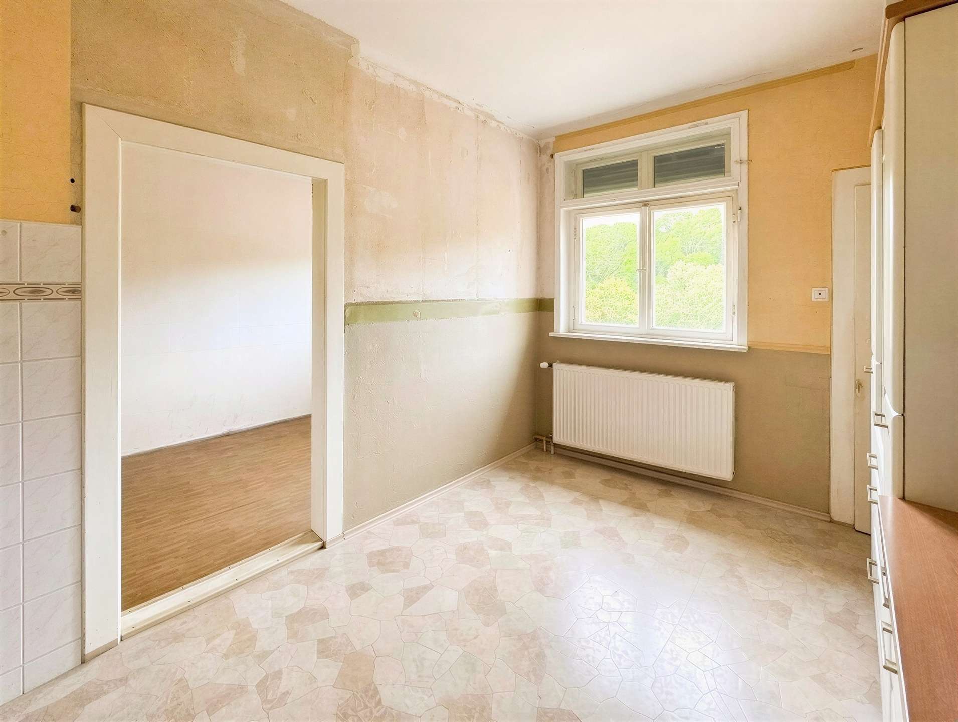 A bright, unfinished kitchen with bare plaster walls, a large window, radiator, and tiled floor; doorway to another room visible on the left.
