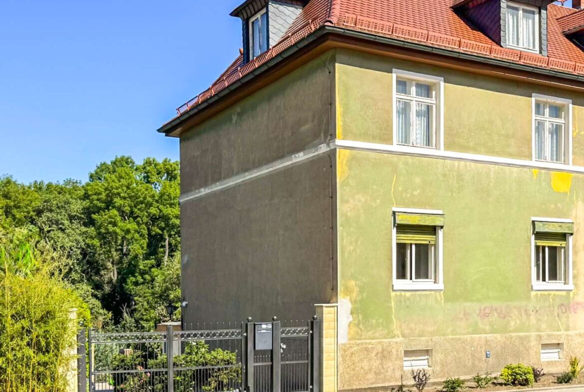 Two-story house with a red tiled roof, pale yellow-green exterior walls, white-framed windows, and a black metal gate in the foreground.