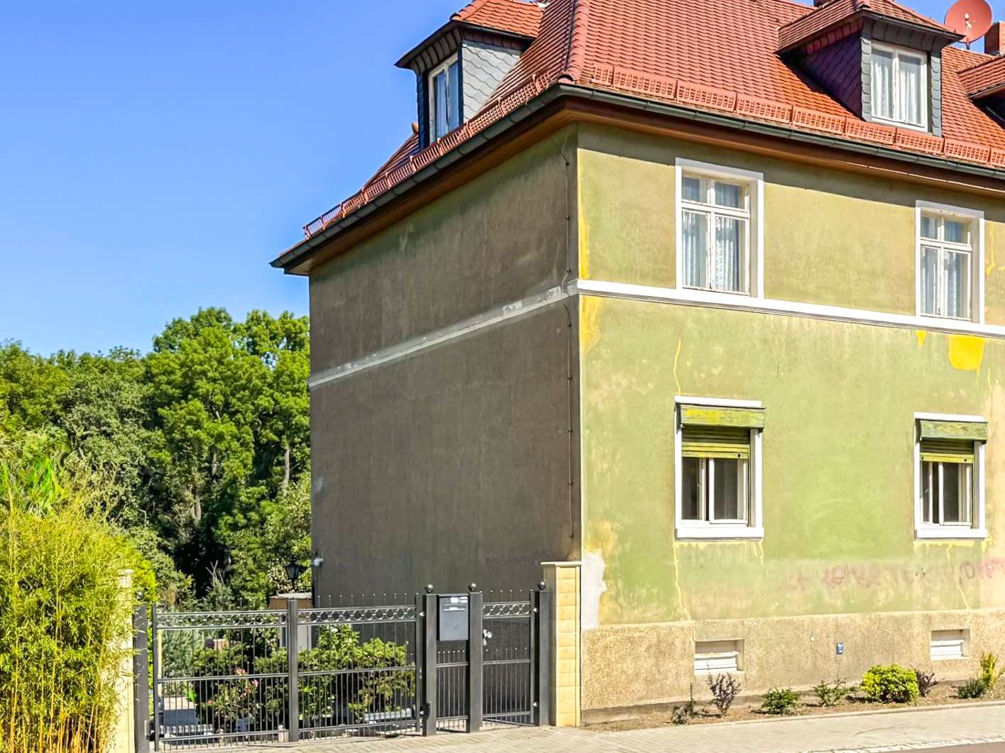 Two-story house with a red tiled roof, pale yellow-green exterior walls, white-framed windows, and a black metal gate in the foreground.