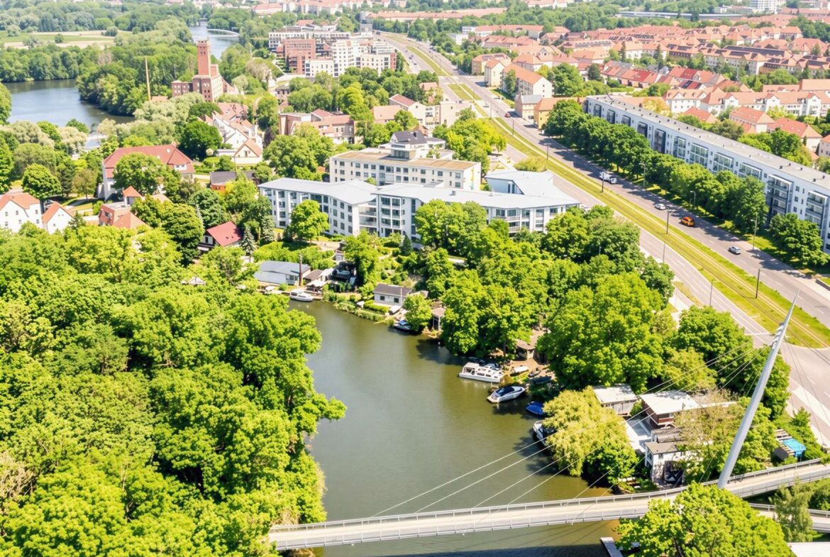 Aerial view of a river winding through a green city with bridges, red-roofed houses, and modern white apartment buildings.