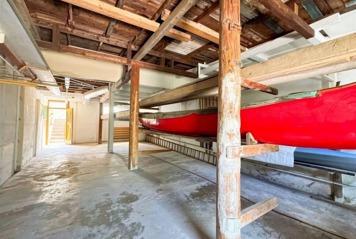 Indoor boat storage with a red canoe on a wooden rack and exposed ceiling beams, ladder nearby.