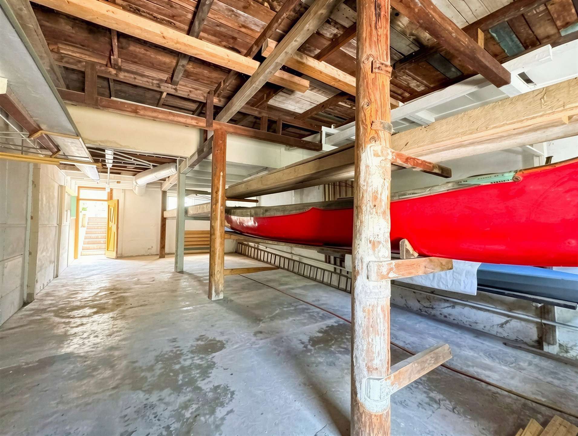 Indoor boat storage with a red canoe on a wooden rack and exposed ceiling beams, ladder nearby.