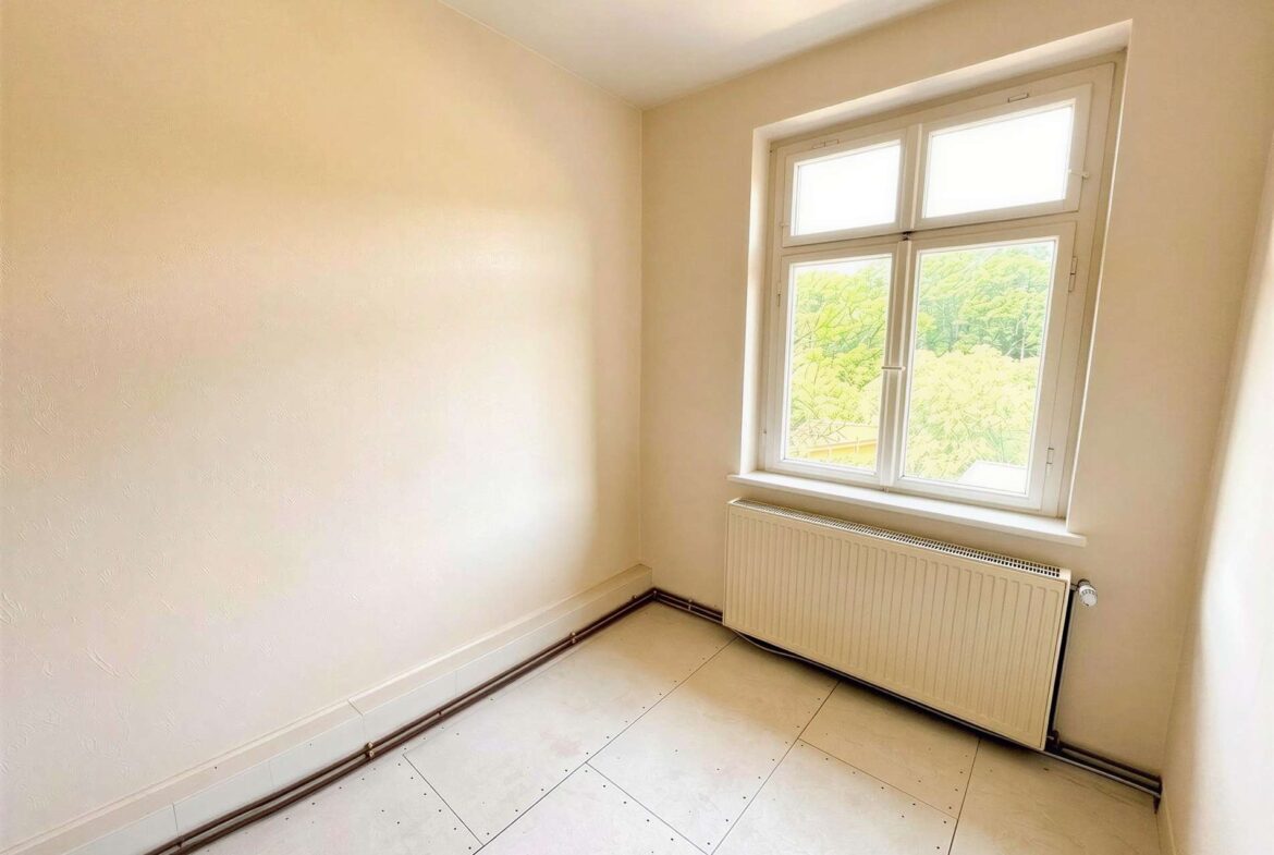 Empty small room with a white-framed window and radiator beneath; pale walls and tiled floor.