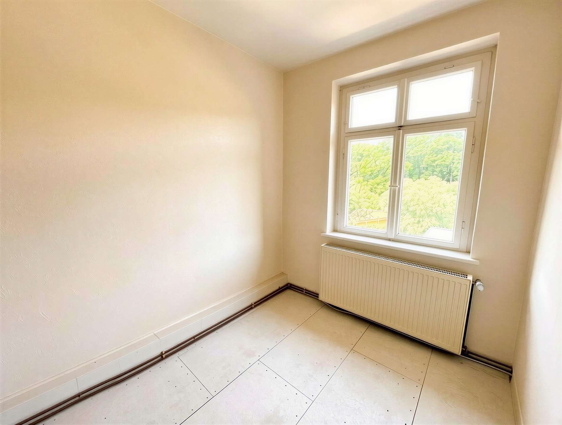 Empty small room with a white-framed window and radiator beneath; pale walls and tiled floor.