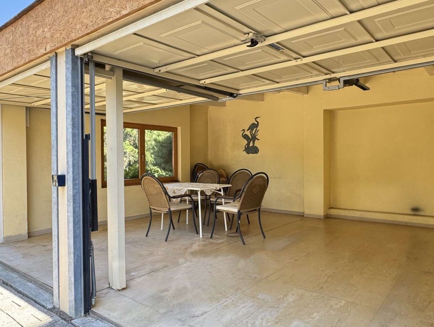 Covered garage dining area with a white table and wicker chairs, yellow walls and a bird mural.