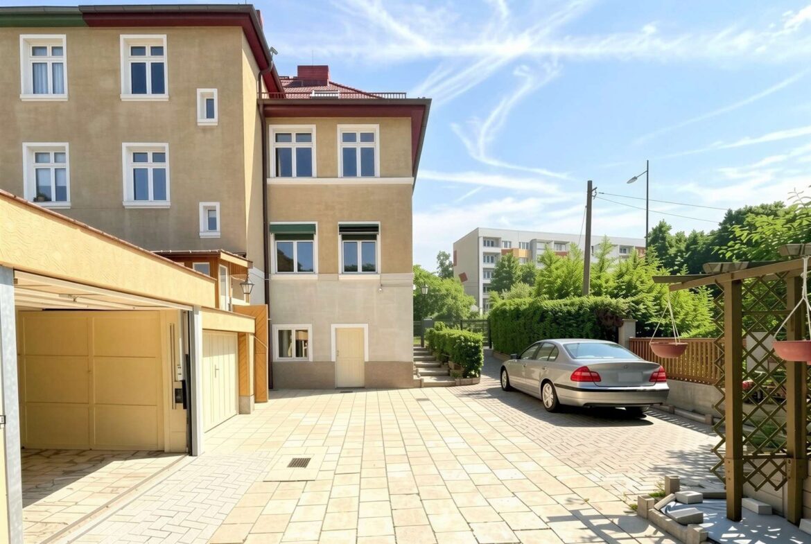 Beige multi-story building with white-framed windows on the left, a tiled courtyard, and a silver car parked near greenery under a blue sky.