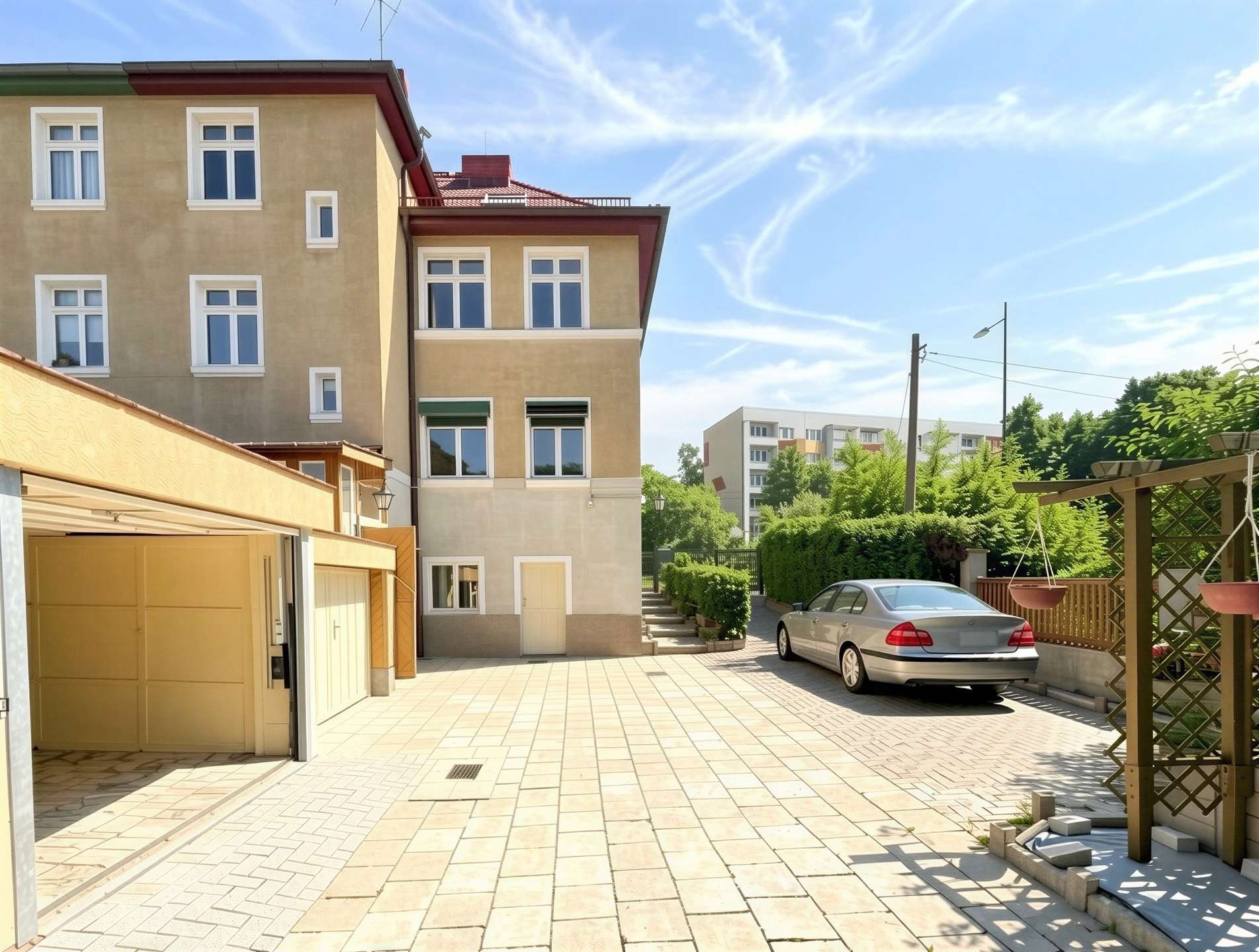 Beige multi-story building with white-framed windows on the left, a tiled courtyard, and a silver car parked near greenery under a blue sky.