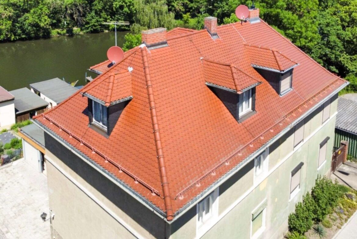 Aerial view of a building with a red-tiled roof, dormer windows and chimneys beside a river and lush green trees.