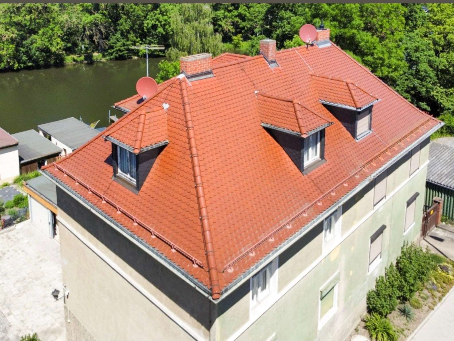 Aerial view of a building with a red-tiled roof, dormer windows and chimneys beside a river and lush green trees.