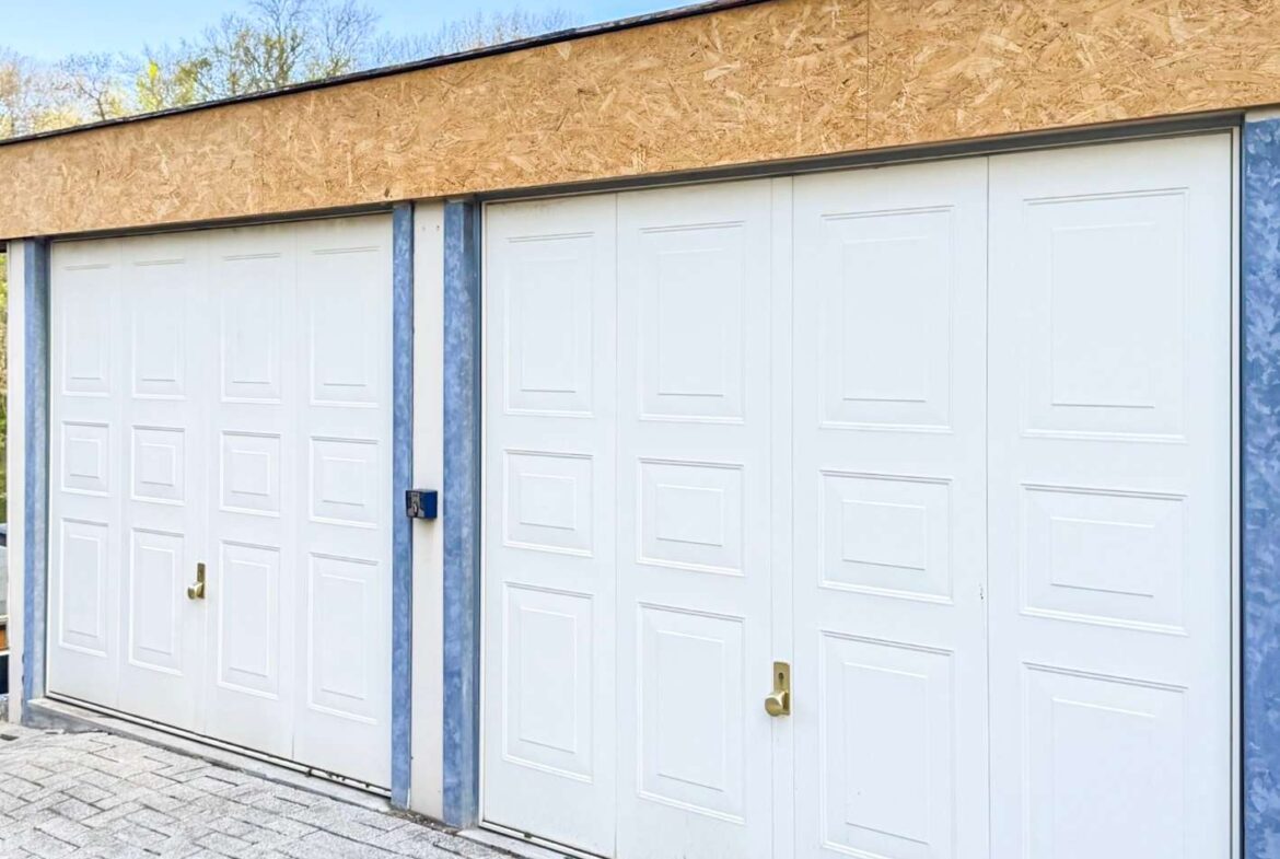 Row of white sectional garage doors with recessed panels, brass handles, and blue metal framing on a paved driveway under a plywood facade top edge.