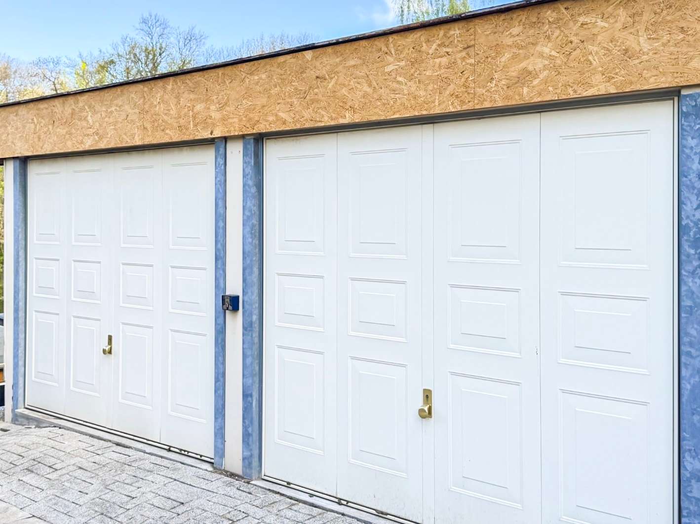 Row of white sectional garage doors with recessed panels, brass handles, and blue metal framing on a paved driveway under a plywood facade top edge.