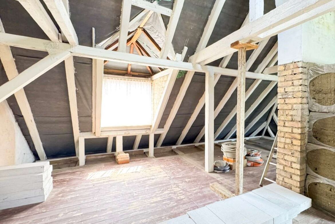 Renovation in a dusty attic with white wooden beams, a small square window, a brick chimney on the right, and coiled cables on the floor.