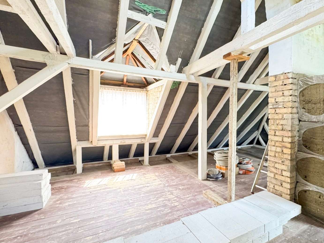 Renovation in a dusty attic with white wooden beams, a small square window, a brick chimney on the right, and coiled cables on the floor.