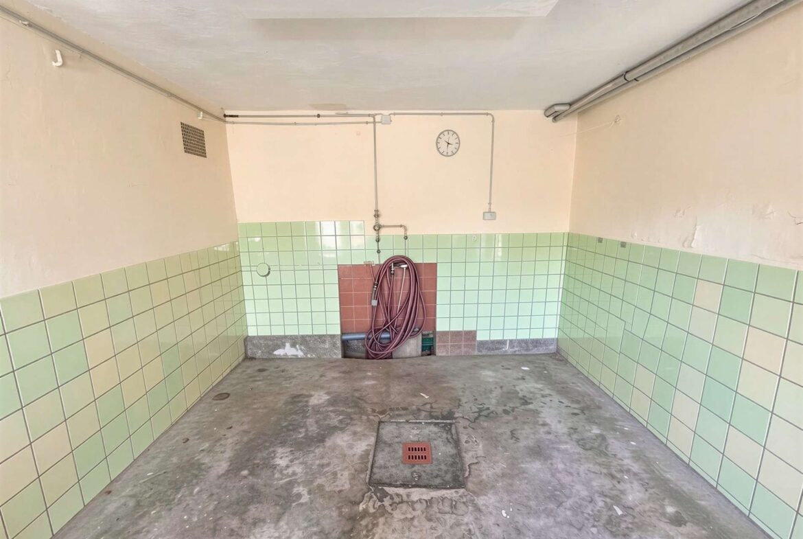 Industrial utility room with green tiled walls, exposed pipes, a coiled hose, and a wall clock above a red-tlo panel center-right; concrete floor with a drain.