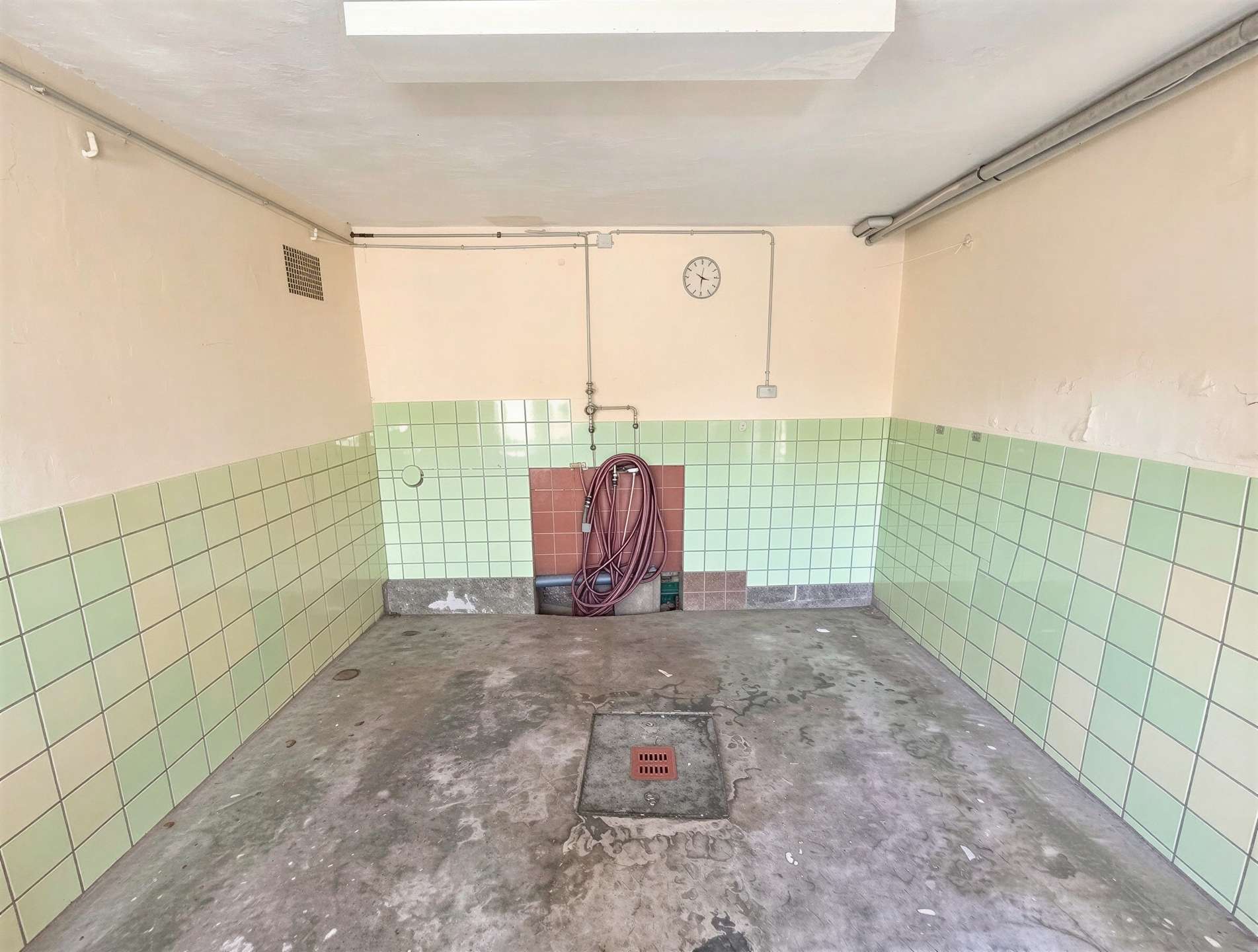 Industrial utility room with green tiled walls, exposed pipes, a coiled hose, and a wall clock above a red-tlo panel center-right; concrete floor with a drain.