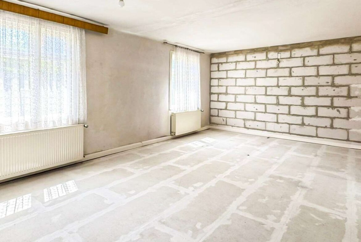 Empty unfinished room with exposed brick wall, bare concrete floor, and two large windows with lace curtains; radiator under one window; single light bulb hanging from ceiling, sunlight streaming in.