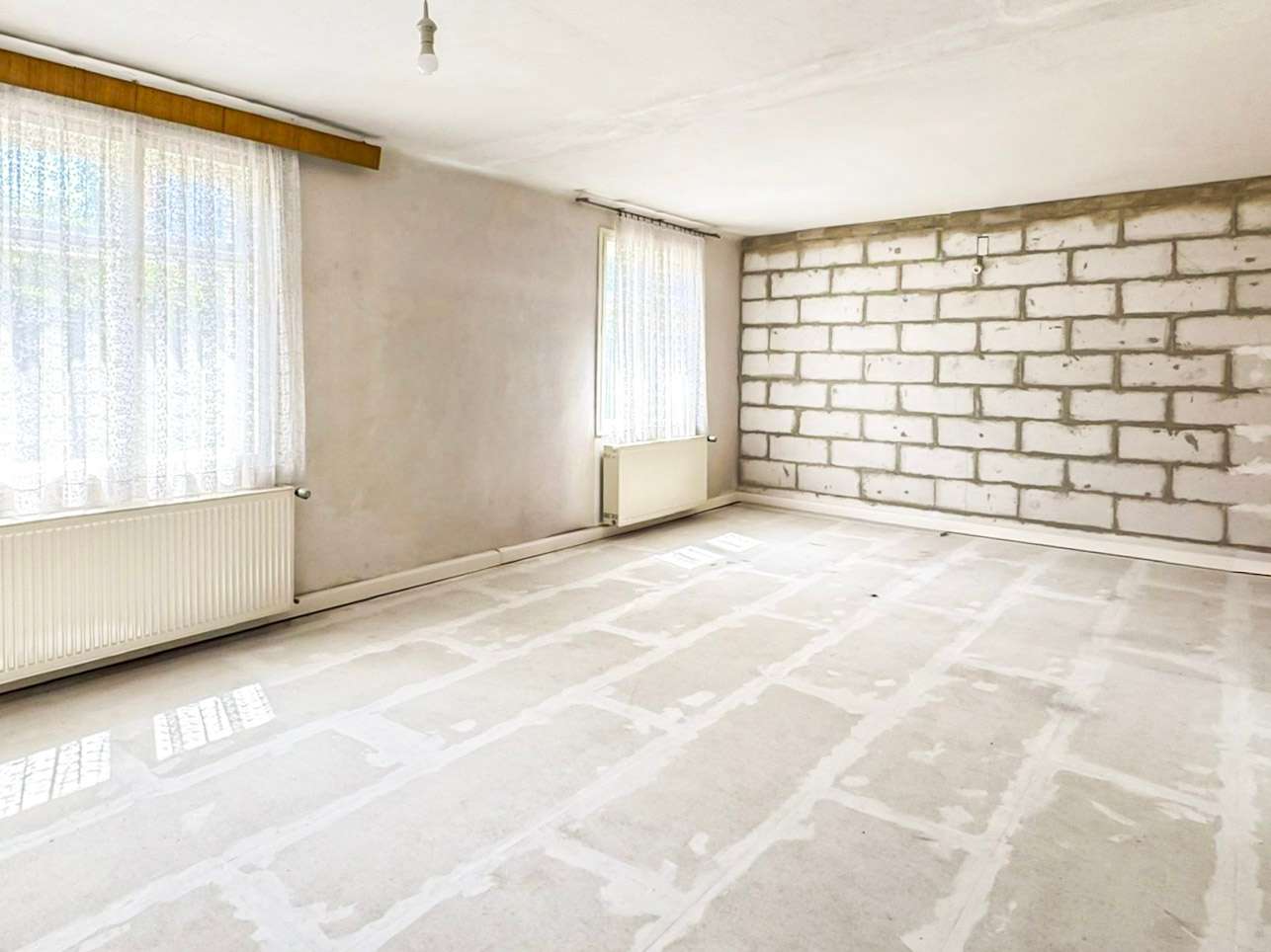 Empty unfinished room with exposed brick wall, bare concrete floor, and two large windows with lace curtains; radiator under one window; single light bulb hanging from ceiling, sunlight streaming in.