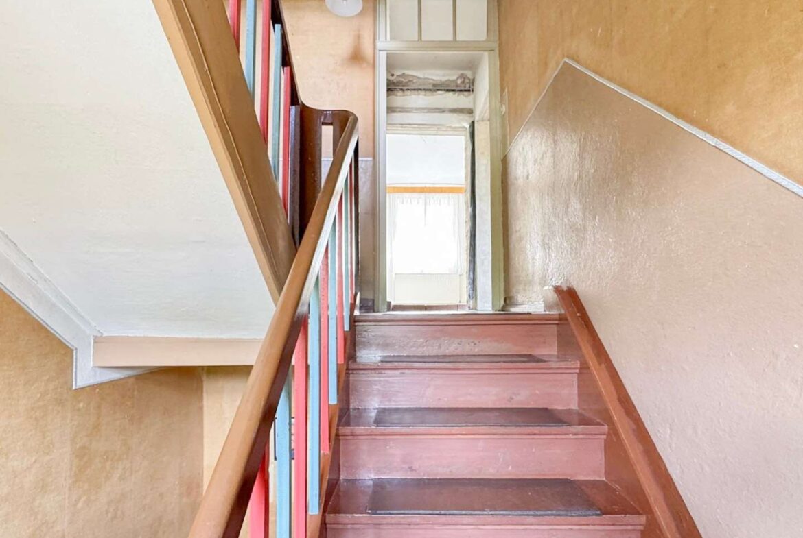 Indoor staircase with pink-brown steps and a colorful blue/red railing, leading to a bright doorway at the top of a worn-warm-toned stairwell, light fixture overhead.
