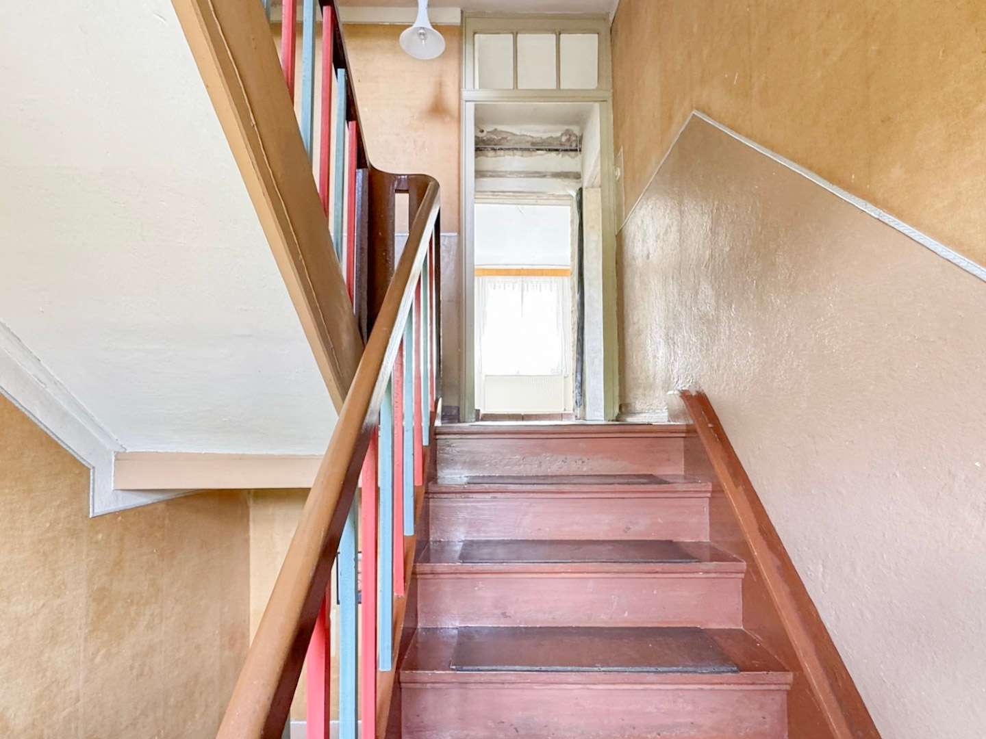 Indoor staircase with pink-brown steps and a colorful blue/red railing, leading to a bright doorway at the top of a worn-warm-toned stairwell, light fixture overhead.