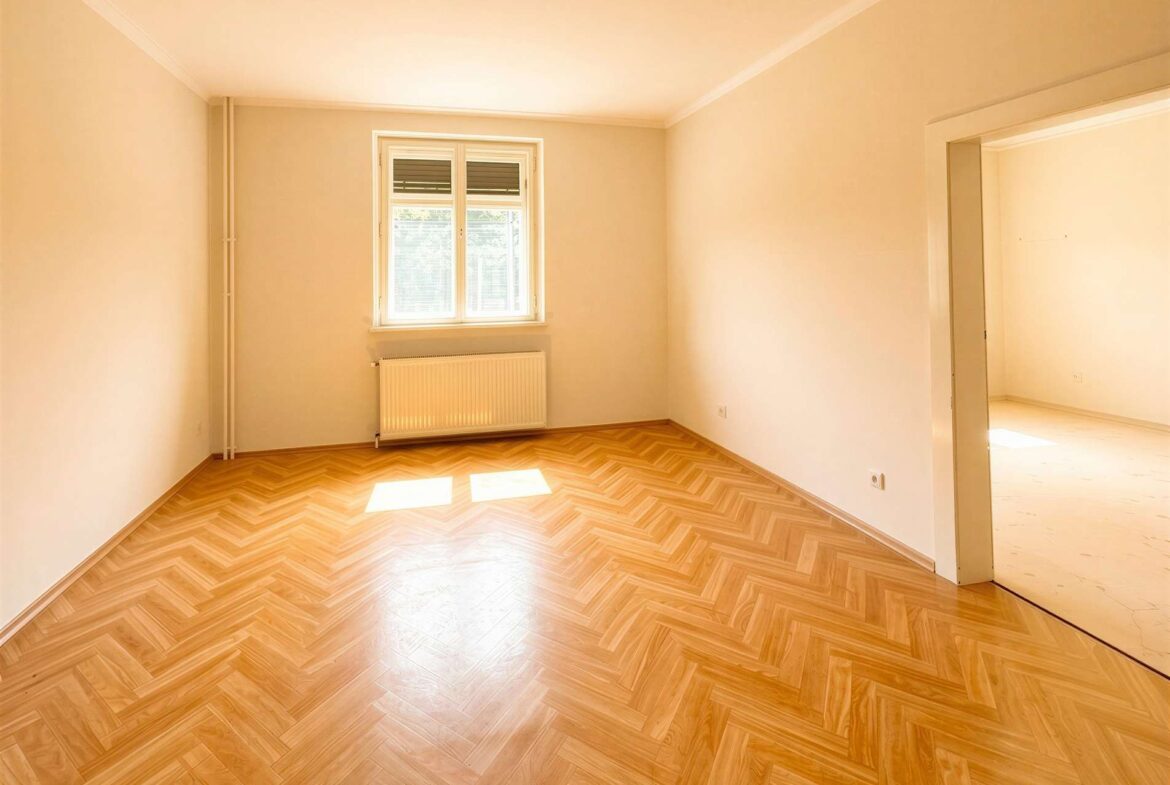 Empty, sunlit room with pale walls, herringbone wood floor, a window with radiator beneath, and an open doorway to another room.