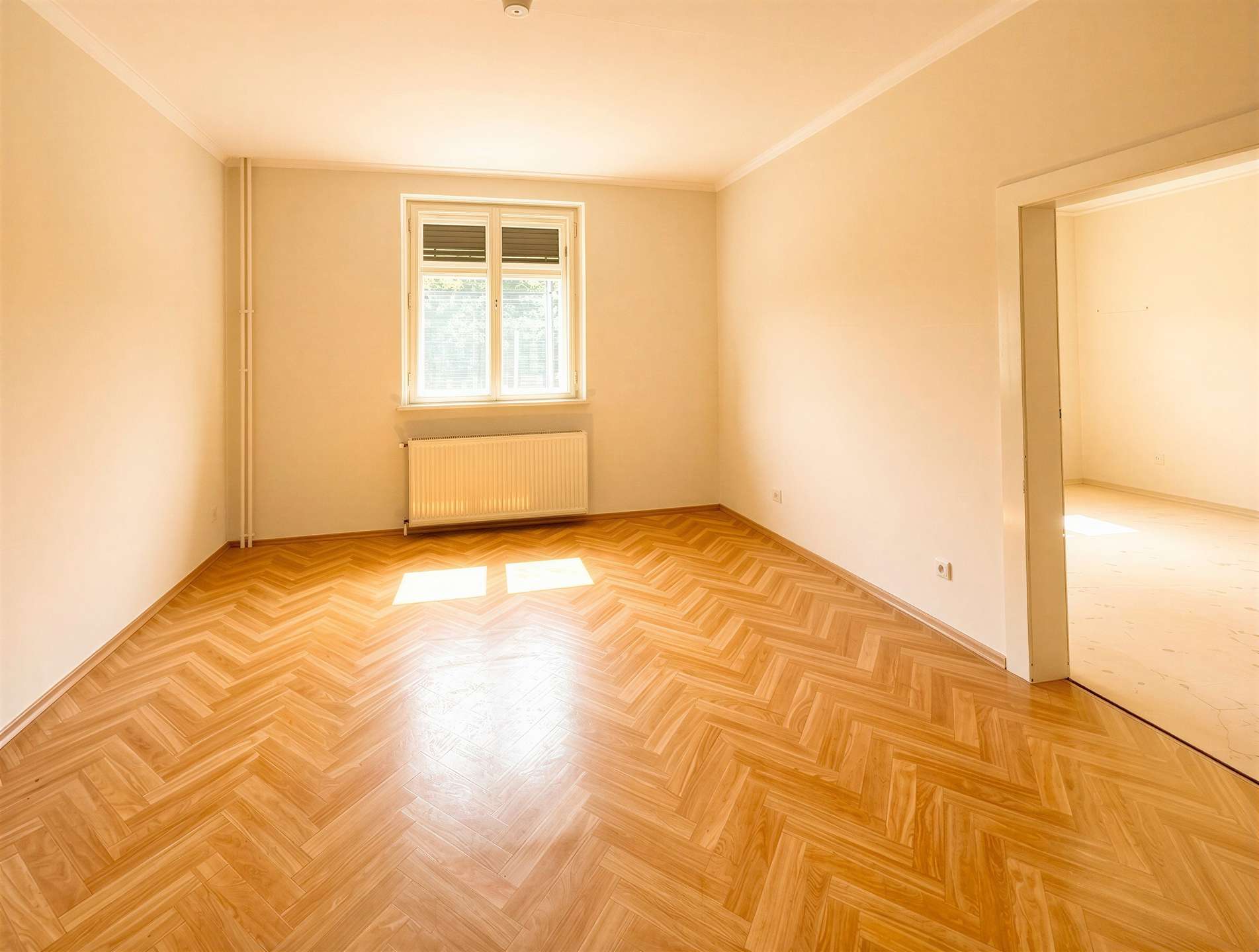 Empty, sunlit room with pale walls, herringbone wood floor, a window with radiator beneath, and an open doorway to another room.