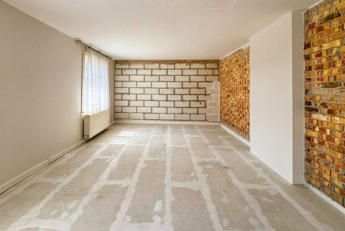 Empty unfinished living room with white painted block walls at the far end, exposed brick wall on the right, and taped protective flooring across the room.
