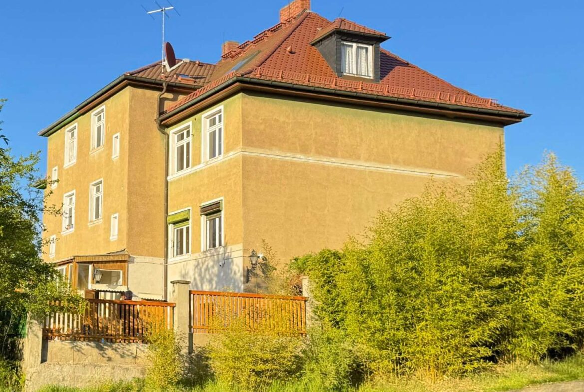 Three-story yellow apartment building with a red tile roof, blue sky, and surrounding greenery.