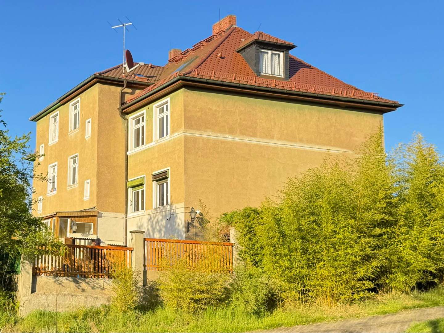 Three-story yellow apartment building with a red tile roof, blue sky, and surrounding greenery.