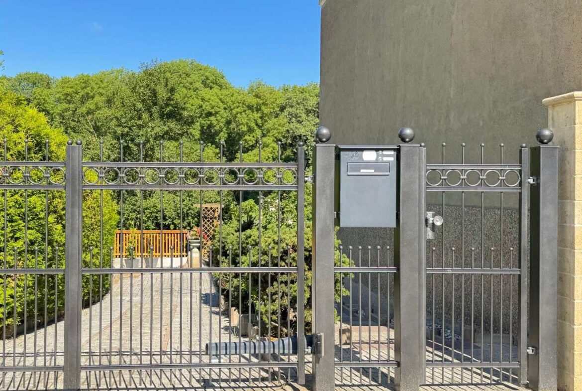 Black iron gate with circular ornaments and a gray mailbox on a pillar; paved path leads to a green garden beyond under a blue sky.