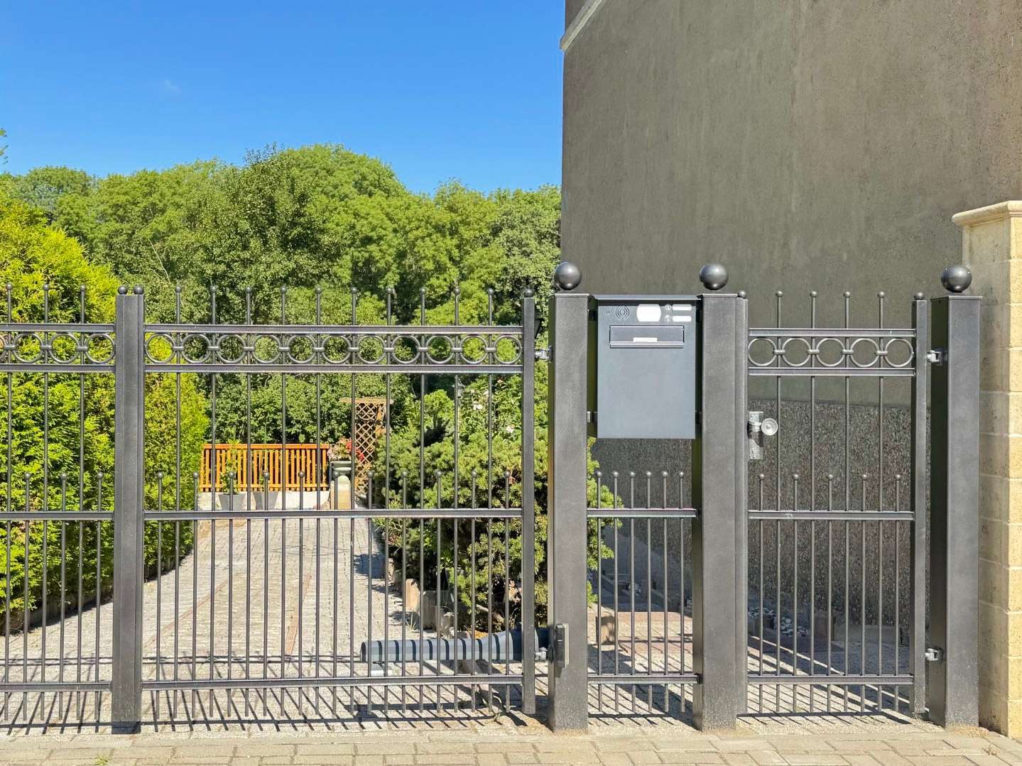 Black iron gate with circular ornaments and a gray mailbox on a pillar; paved path leads to a green garden beyond under a blue sky.