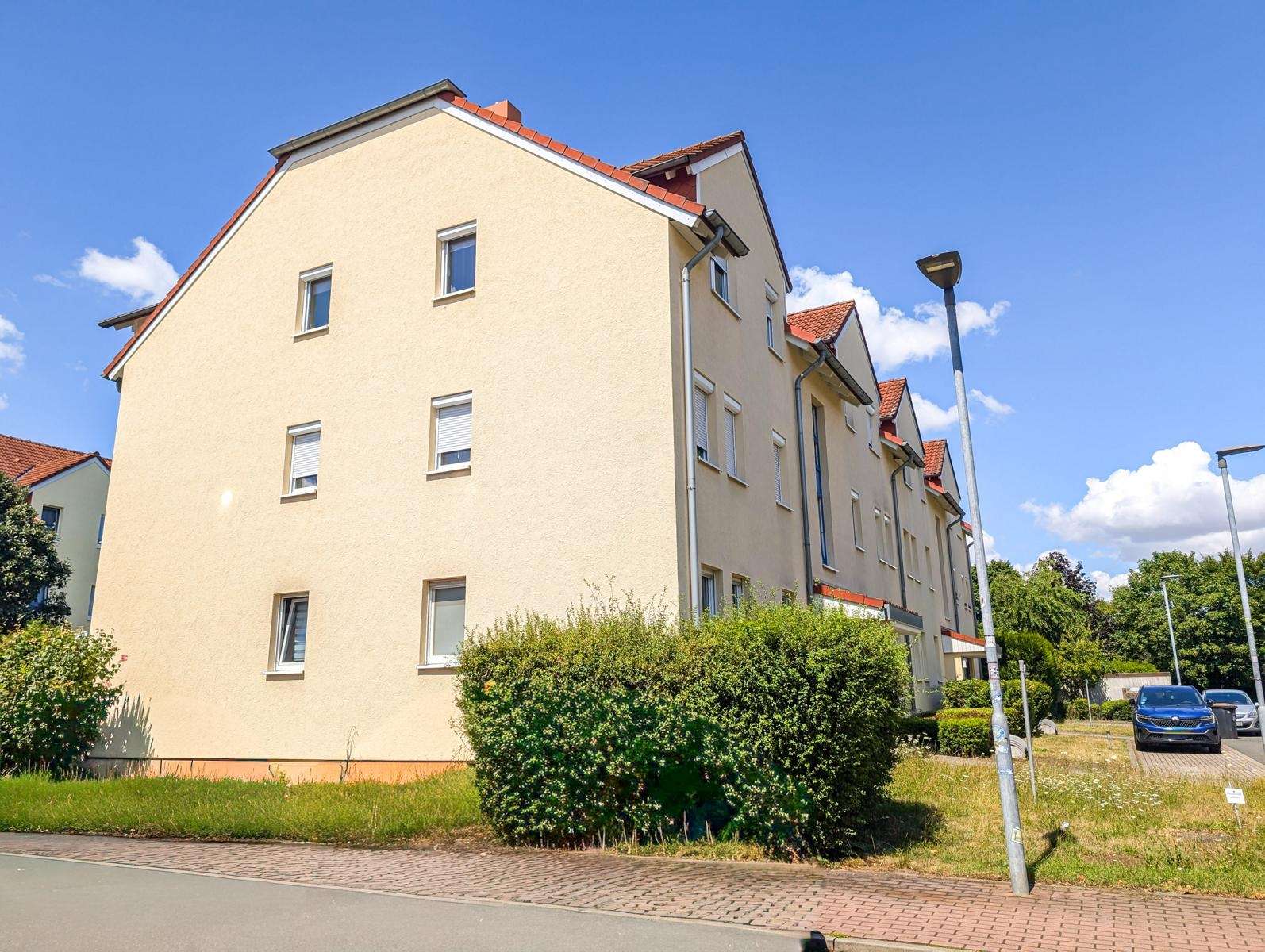 Three-story beige apartment building with a red-tiled roof, along a sunny street with hedges and a parked blue car.
