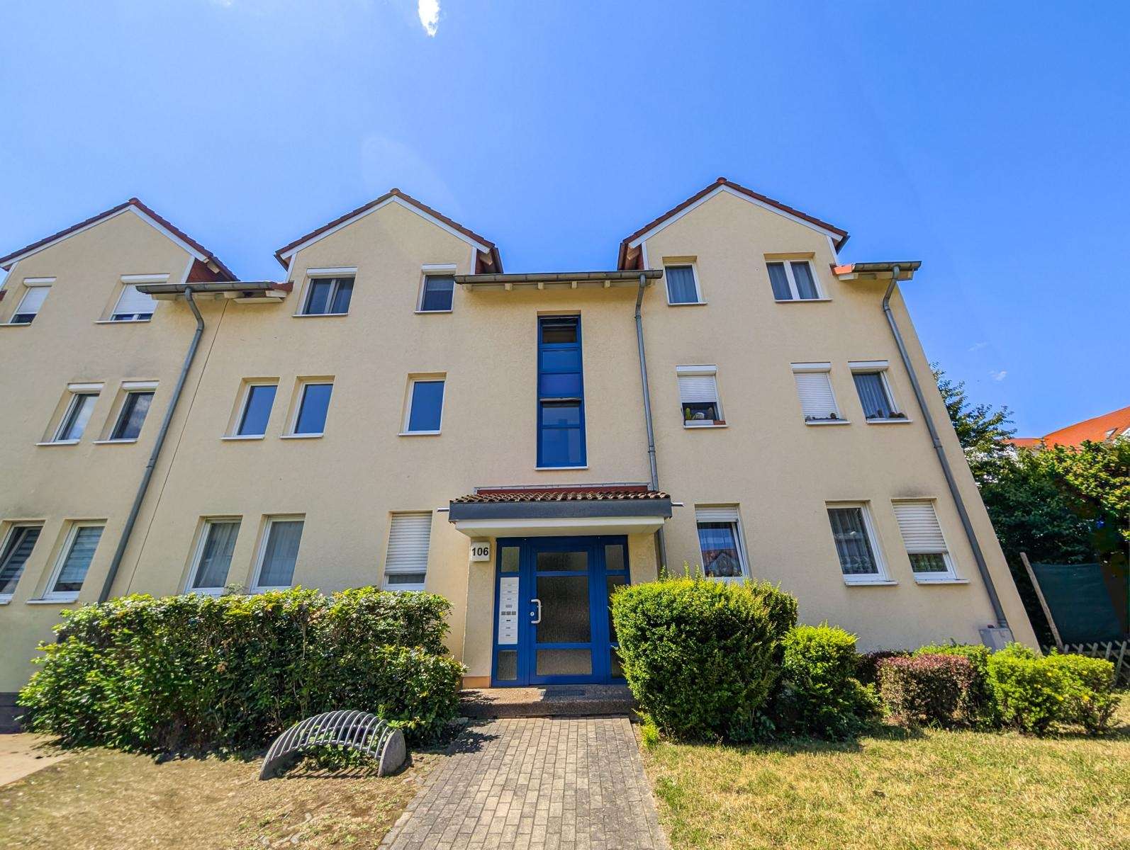 Beige three-story apartment building with a blue central entrance door, many white-framed windows, and a clear blue sky.
