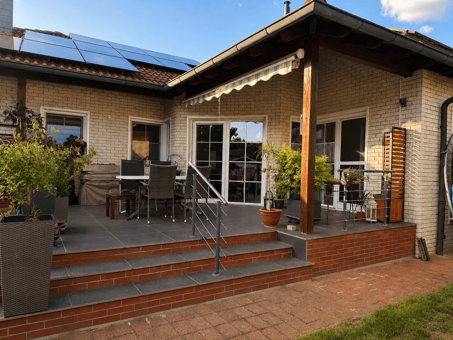 Brick house with a tiled patio, solar panels on the roof, and an outdoor dining area with a table, chairs, and potted plants on a raised deck under an awning.