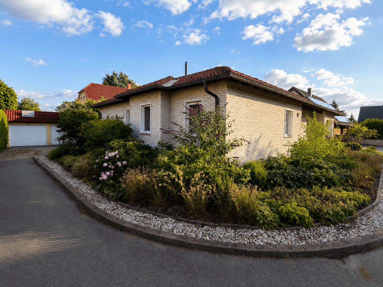 Single-story brick house with a red tile roof, surrounded by a lush garden and a curved driveway.