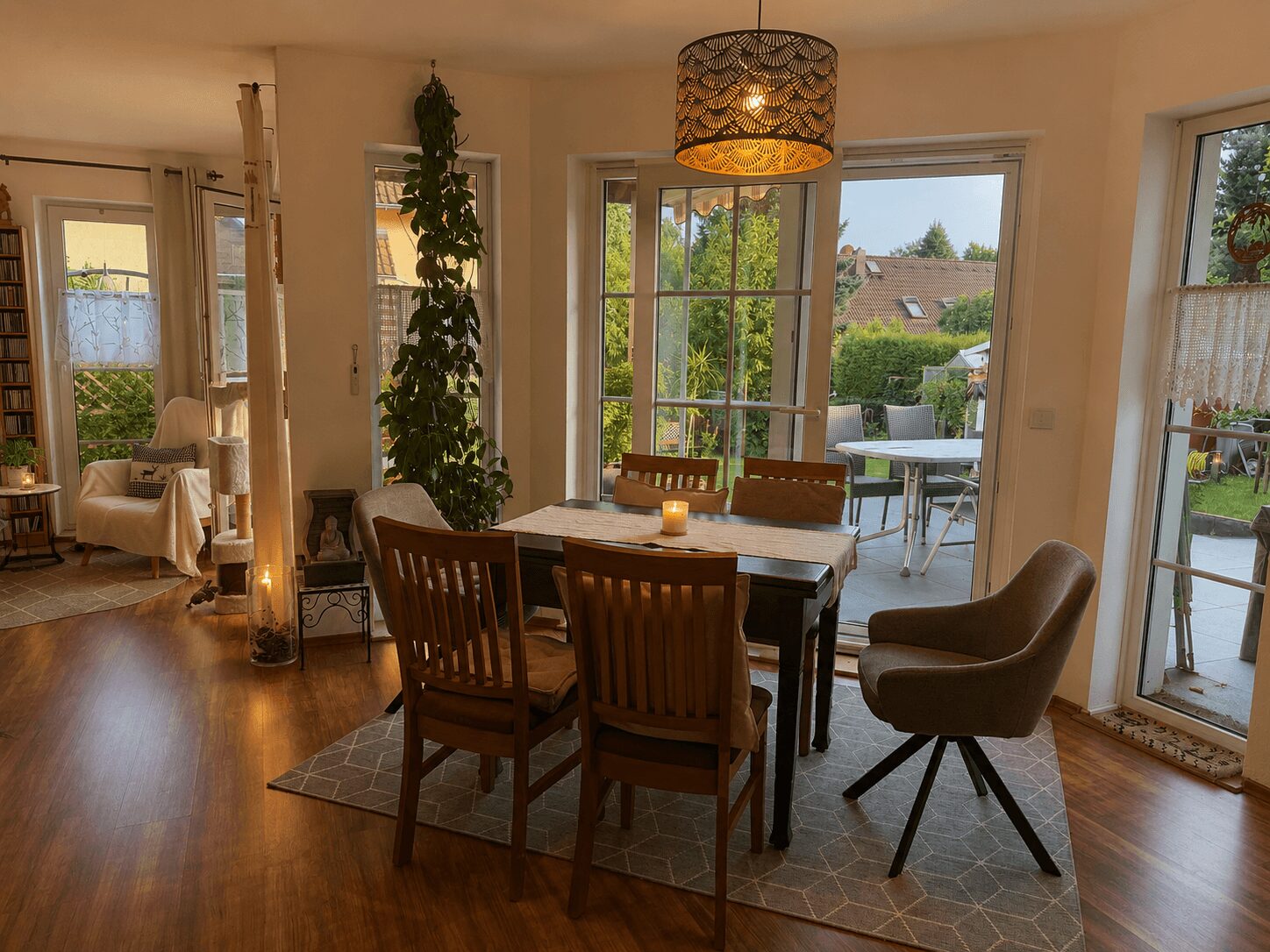 Bright dining area with a wooden table and six chairs, lit by a candle and a hanging decorative lantern, adjacent to large windows and a glass door to a patio garden.