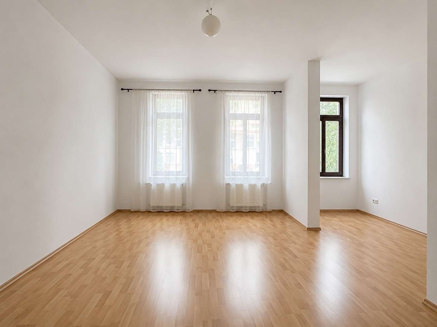 Bright empty living room with light wood floor, white walls, and two large windows with sheer curtains.