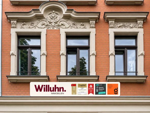 Brick building façade with ornate stone decorations above three tall windows and a Willuhn Immobilien sign with award stickers below.