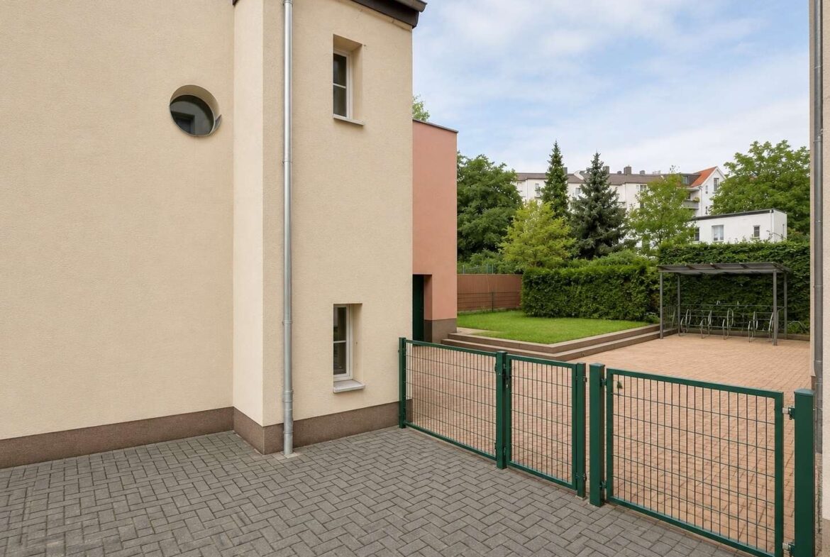 Exterior courtyard with beige building, green metal gate, and a bike shelter on a paved area.