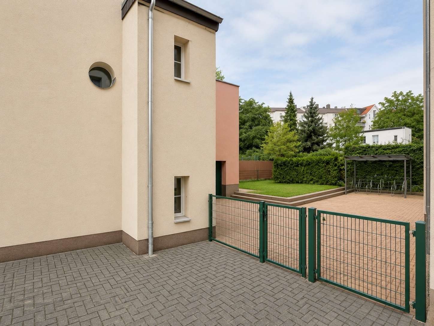 Exterior courtyard with beige building, green metal gate, and a bike shelter on a paved area.