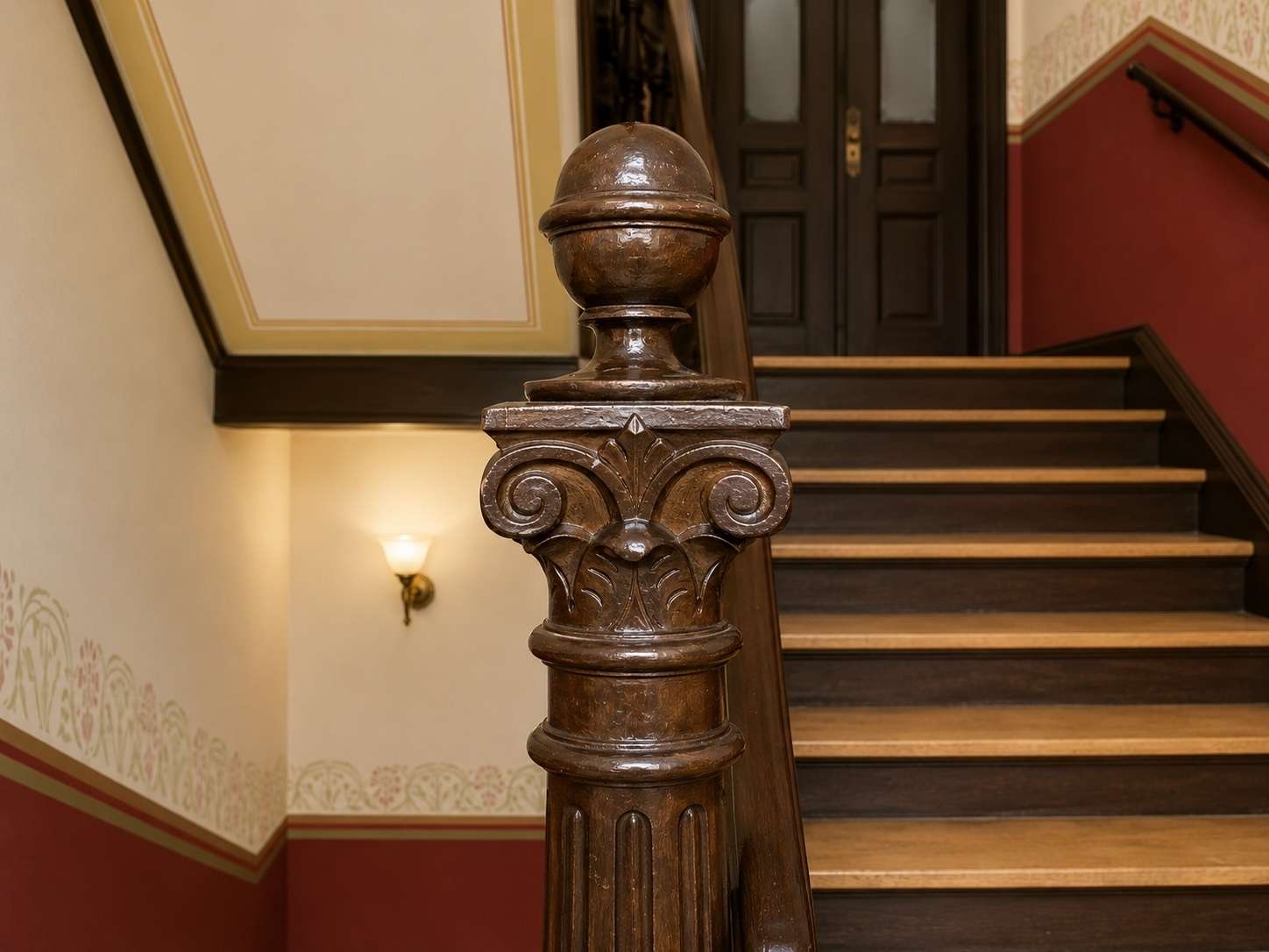 Ornate wooden staircase newel post with carved scrolls and a rounded cap, in a warmly lit interior stairway.