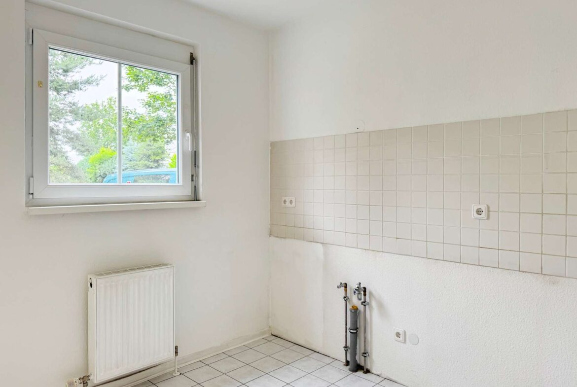 Bright empty kitchen area with a white-framed window, radiator on the left, tiled wall to the right, and exposed plumbing at the base.