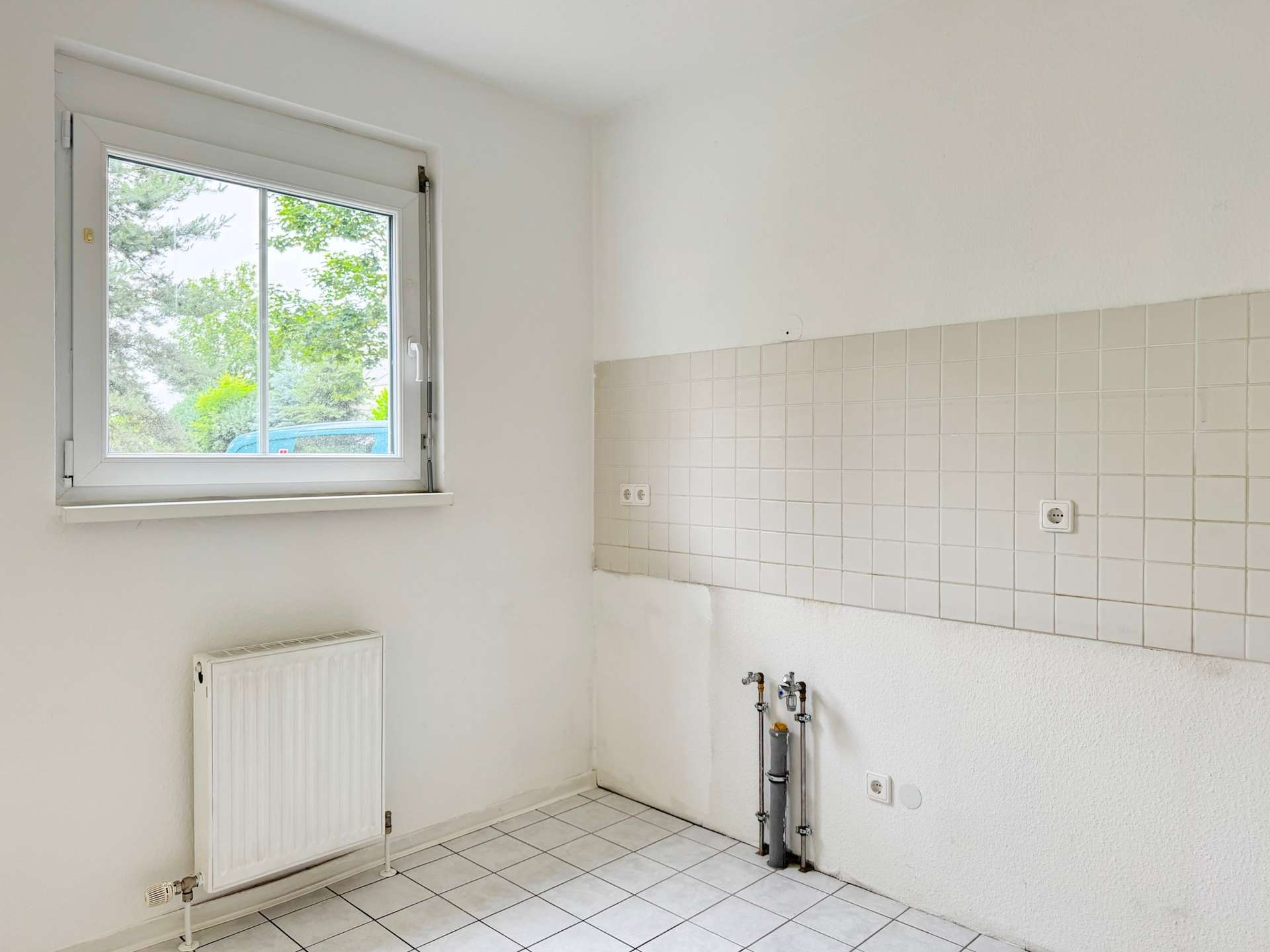 Bright empty kitchen area with a white-framed window, radiator on the left, tiled wall to the right, and exposed plumbing at the base.