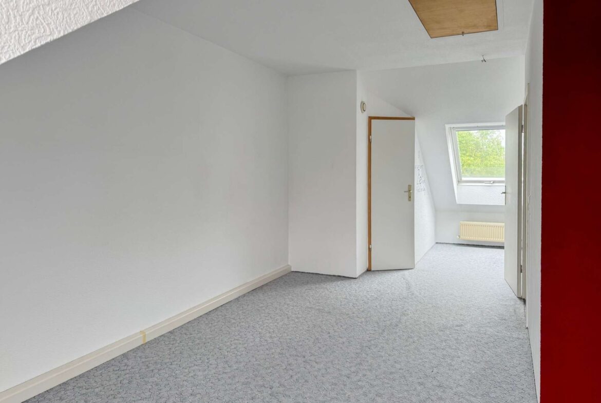 Empty white hallway with a sloped ceiling, open door, and a window letting in daylight; pale patterned carpet and radiator visible.
